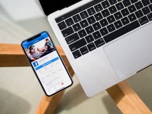 Facebook page displayed on mobile phone beside MacBook Pro 